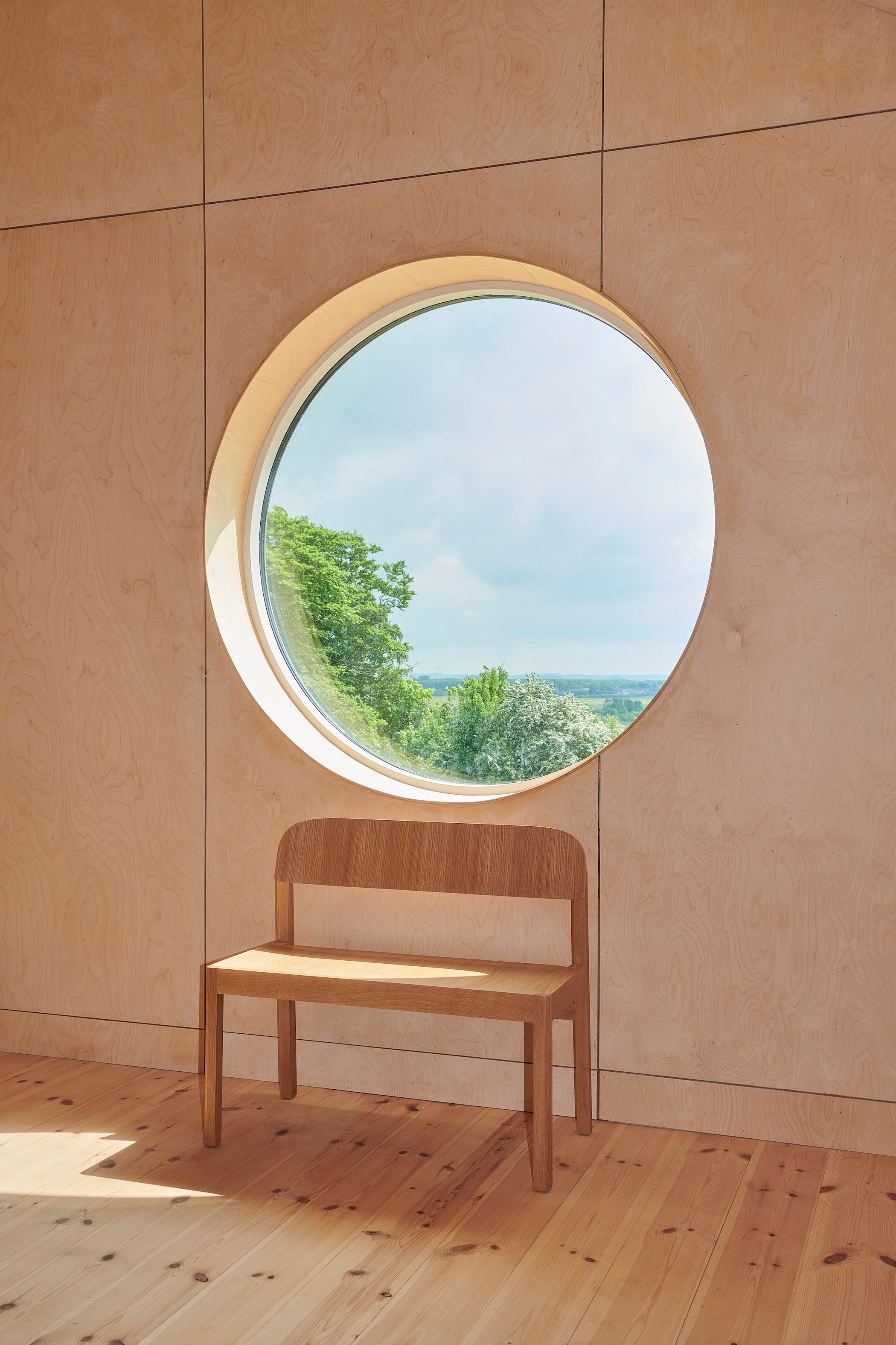 Wooden bench in front of a round window with a view of greenery on a beige wall.