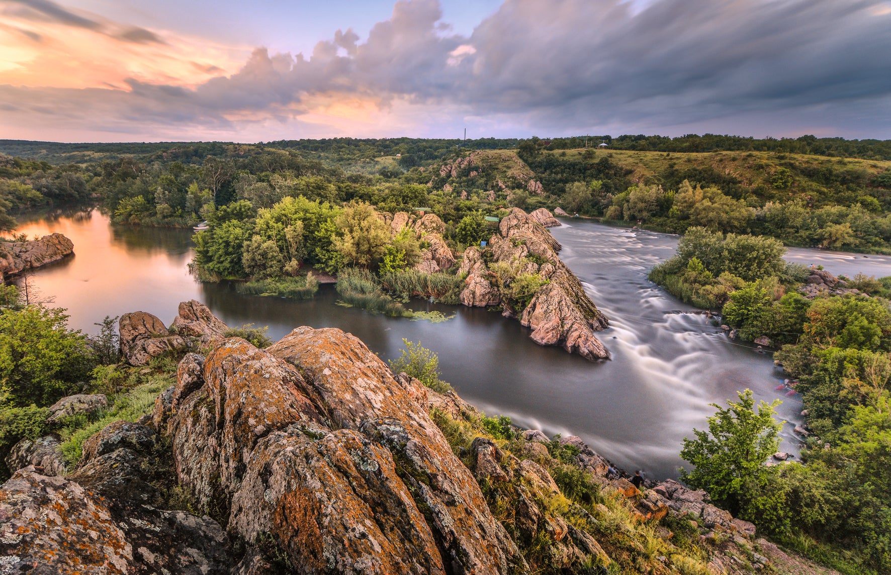 Scenic view of a river with rocky formations and greenery under a colorful sky.